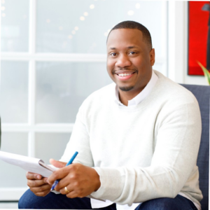 Mychal smiles in white sweater holding pen and notepad for a headshot.